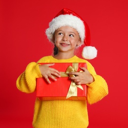 Photo of Cute little girl in Santa hat with Christmas gift on red background. Space for text