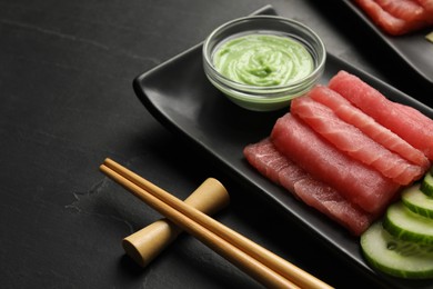 Photo of Tasty sashimi (pieces of fresh raw tuna), cucumber slices and wasabi sauce on black plate, closeup