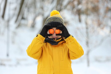Cute little boy covering eyes with tangerines in snowy park on winter day