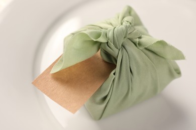 Photo of Furoshiki technique. Gift packed in green fabric with blank card on plate, closeup