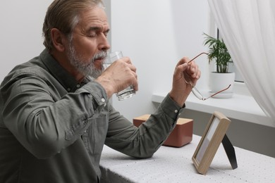 Photo of Upset senior man drinking glass of water near photo frame on table at home. Loneliness concept