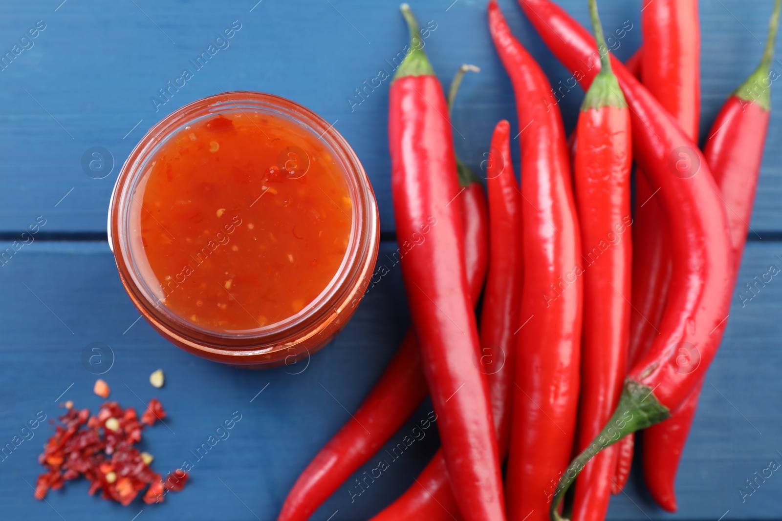 Photo of Spicy chili sauce in jar and peppers on blue wooden table, flat lay