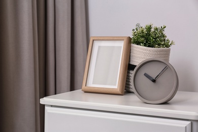 Stylish analog clock, photo frame and potted plant on chest of drawers in room. Space for text