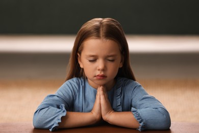 Photo of Cute little girl with hands clasped together praying at table indoors