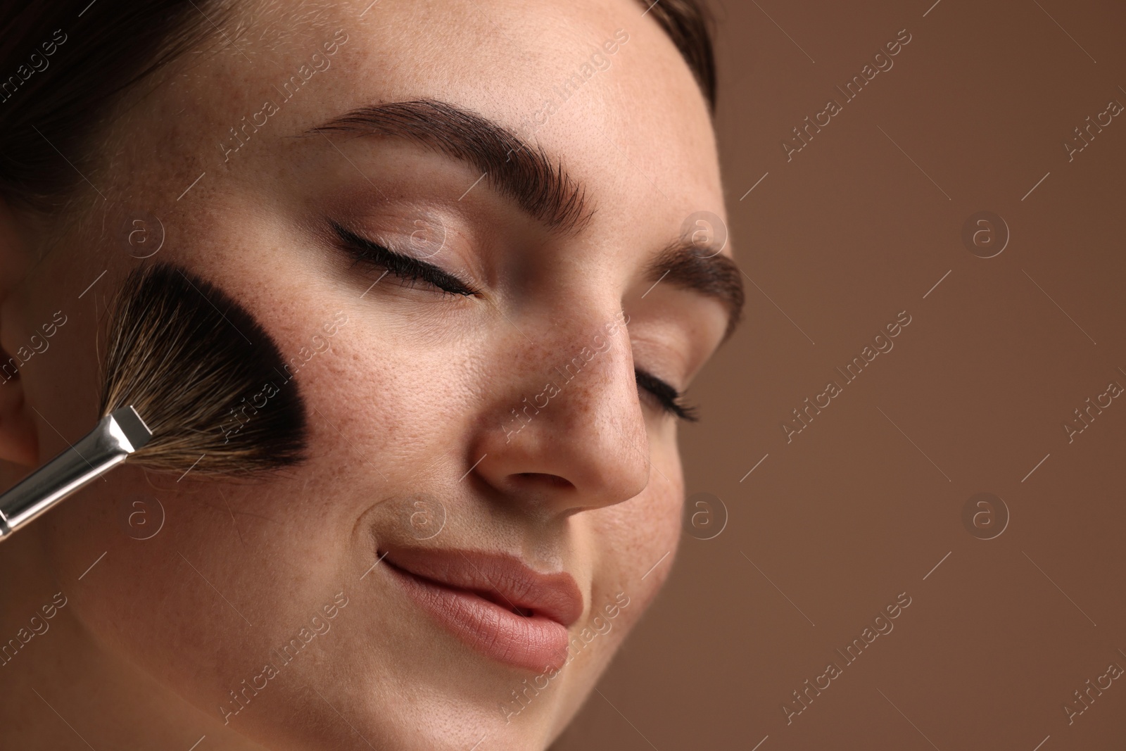 Photo of Beautiful woman with freckles applying makeup with brush on brown background, closeup