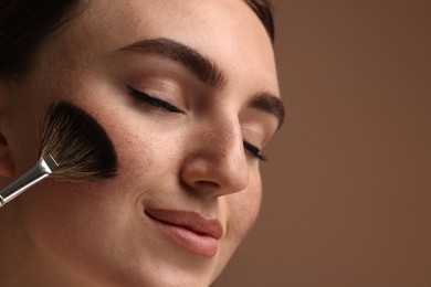 Beautiful woman with freckles applying makeup with brush on brown background, closeup