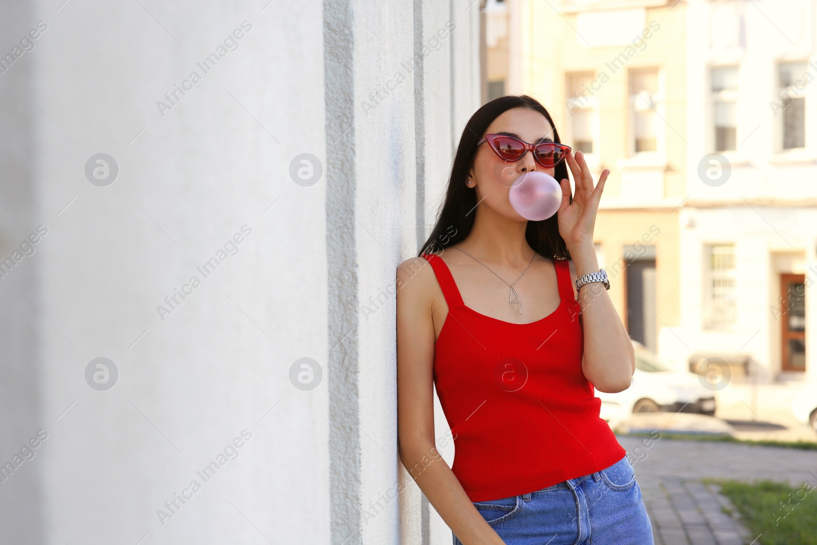 Photo of Beautiful woman in stylish sunglasses blowing gum near building outdoors, space for text