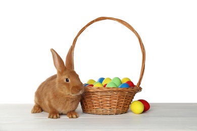 Photo of Cute bunny and basket with Easter eggs on table against white background