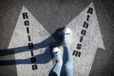 Image of Philosophical or religious position. Woman standing on road marking arrow with word Atheism, closeup