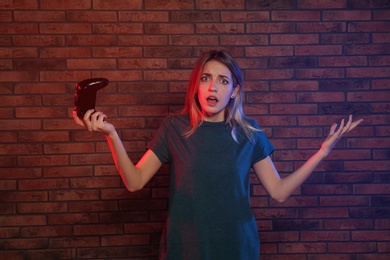 Emotional young woman playing video games with controller near brick wall