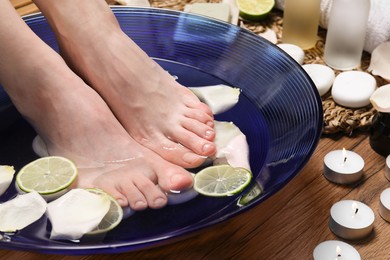 Photo of Woman soaking her feet in bowl with water, petals and lime slices on wooden floor, closeup. Spa treatment