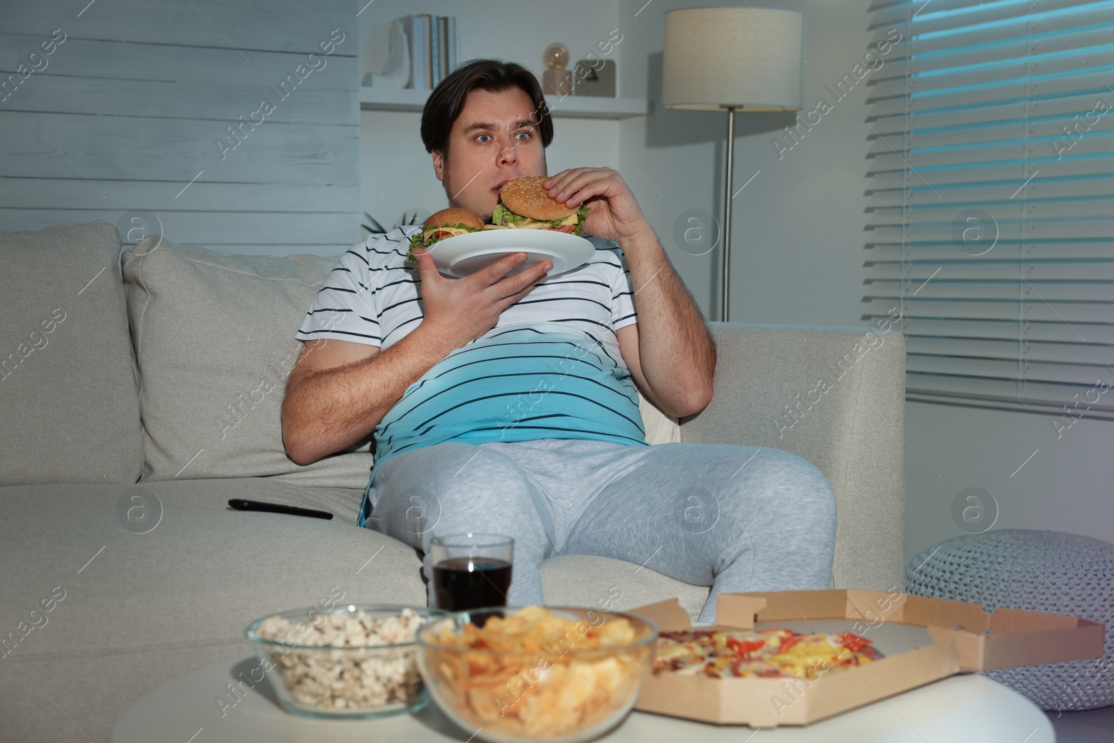 Photo of Overweight man with plate of burgers and French fries on sofa at home