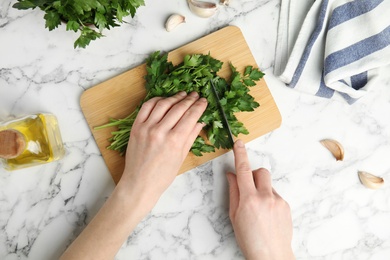 Photo of Woman cutting fresh green parsley on wooden board at marble table, top view