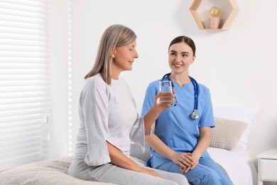 Photo of Senior woman with glass of water and young healthcare worker sitting on bed indoors