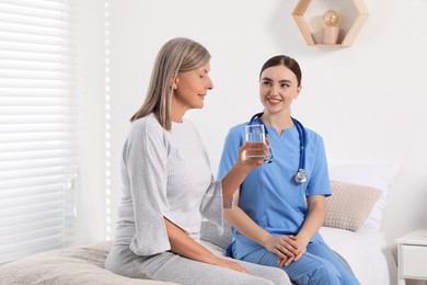 Senior woman with glass of water and young healthcare worker sitting on bed indoors