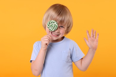 Photo of Happy little boy covering his eye with bright lollipop swirl on orange background