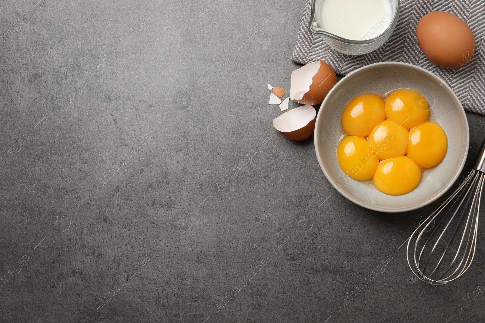 Photo of Bowl with raw egg yolks, whisk and jug of milk on grey table, flat lay. Space for text