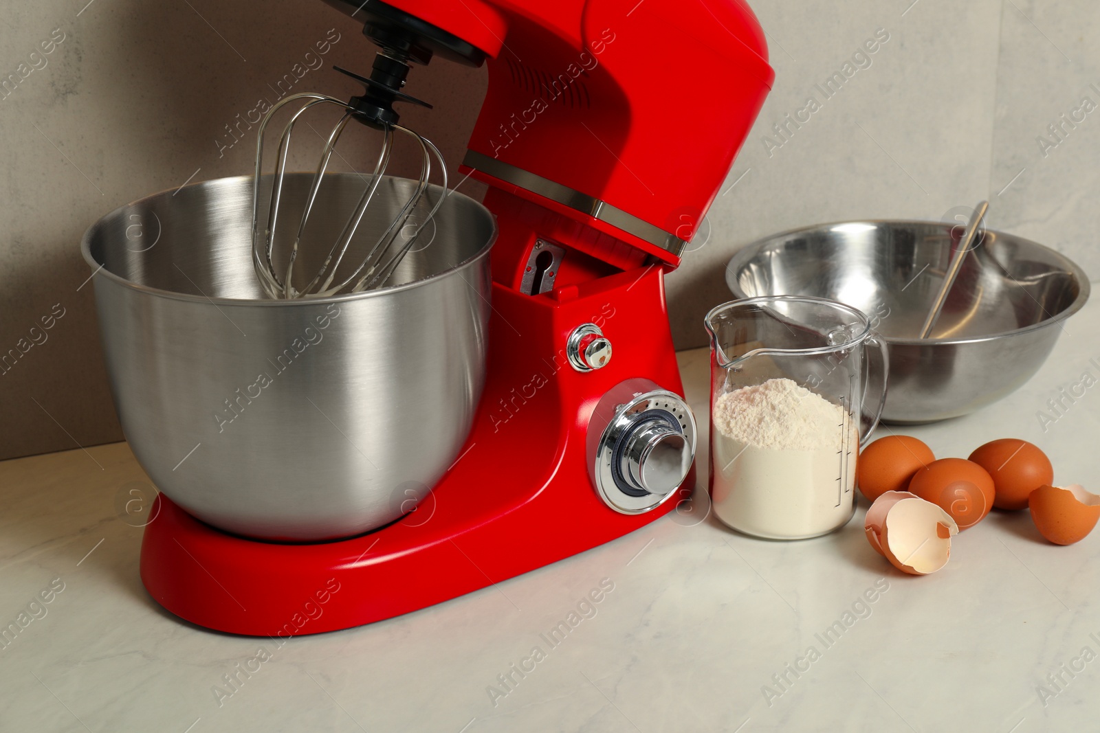 Photo of Modern red stand mixer, eggs, container with flour and bowl on white marble table