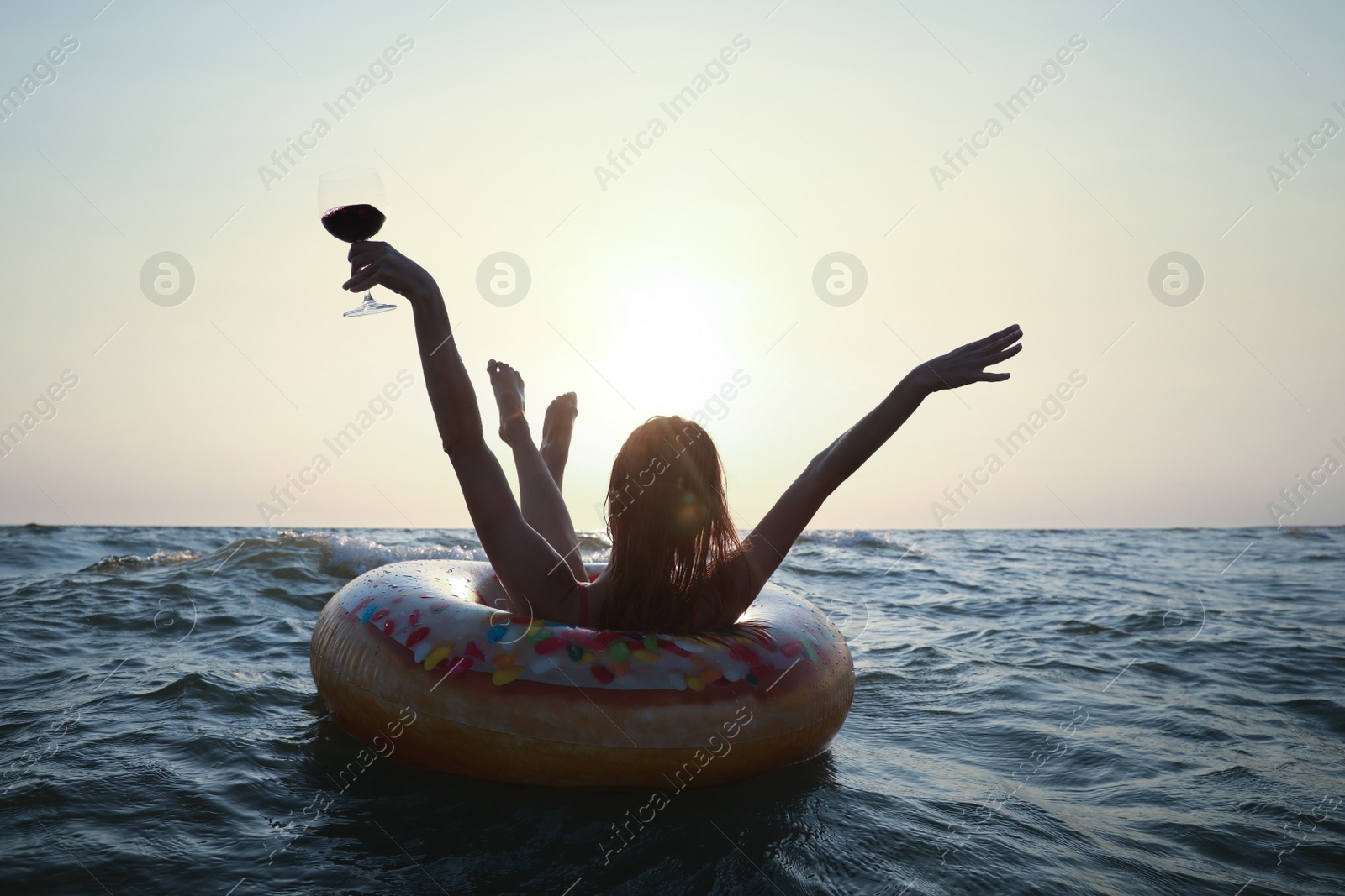 Photo of Happy woman with glass of wine and inflatable ring in sea at sunset, back view