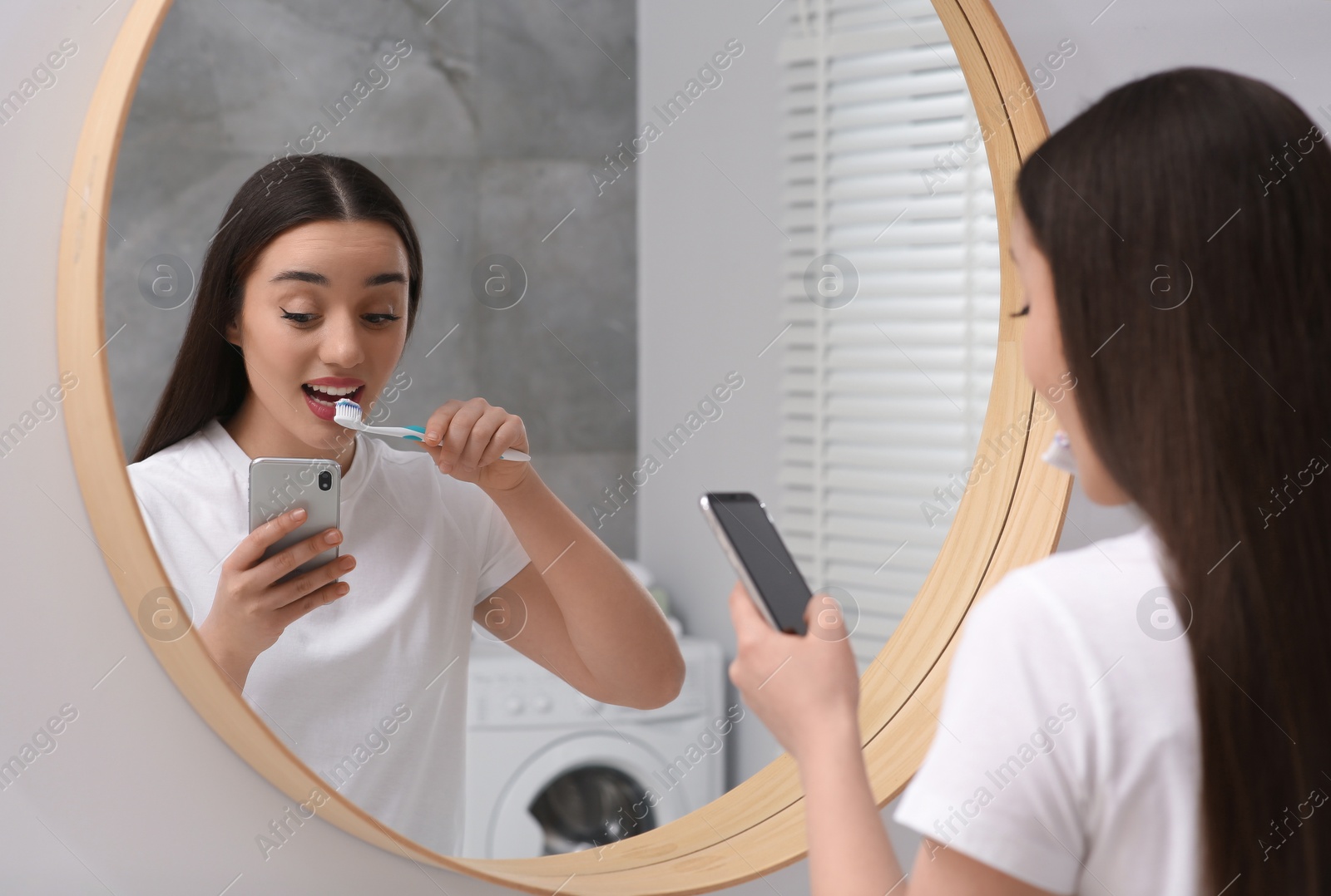 Photo of Beautiful young woman using smartphone while brushing teeth in bathroom. Internet addiction