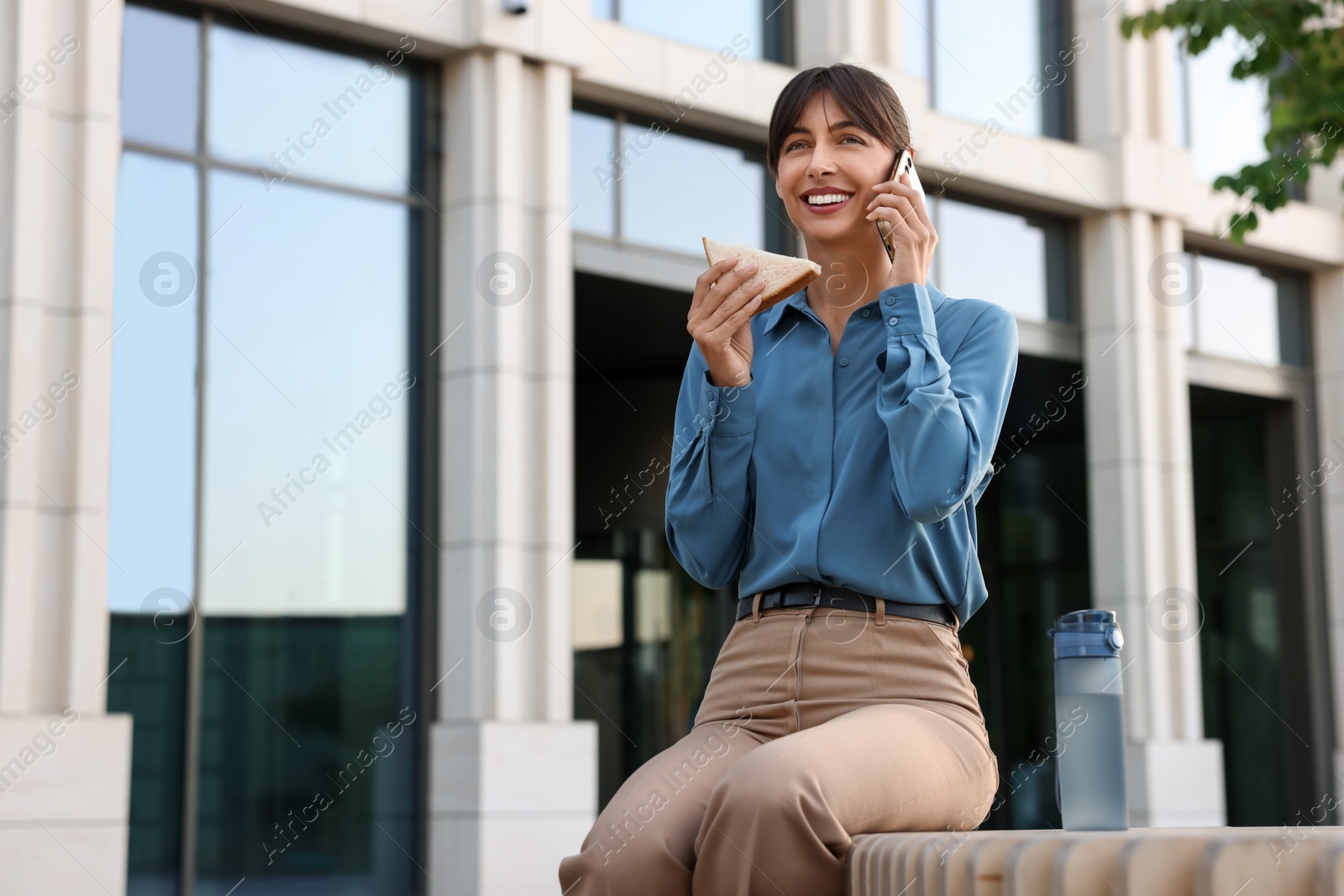 Photo of Happy businesswoman with sandwich talking on smartphone during lunch on bench outdoors, low angle view