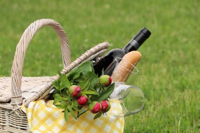 Photo of Picnic basket with wine, bread and flowers on green grass outdoors, closeup