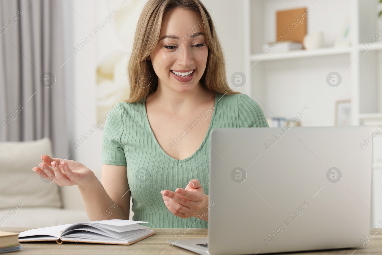 Photo of Young woman waving hello during video chat via laptop at wooden table indoors