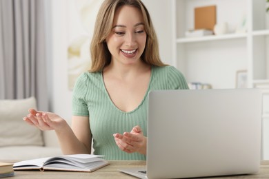 Photo of Young woman waving hello during video chat via laptop at wooden table indoors