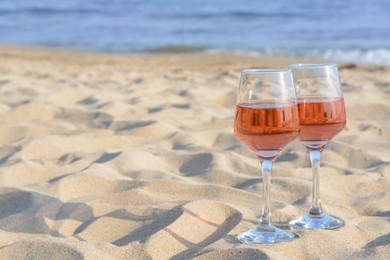 Glasses of tasty rose wine on sand near sea, space for text