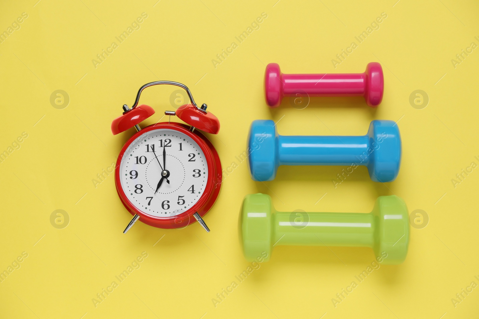 Photo of Alarm clock and dumbbells on yellow background, flat lay. Morning exercise