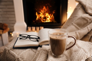 Cup of coffee, book and glasses near fireplace indoors, space for text. Cozy atmosphere
