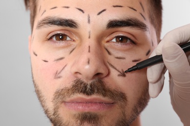 Doctor drawing marks on man's face for cosmetic surgery operation against grey background, closeup