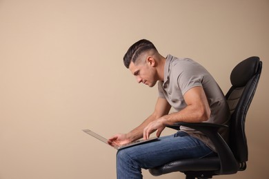 Man with poor posture using laptop while sitting on chair against beige background, space for text