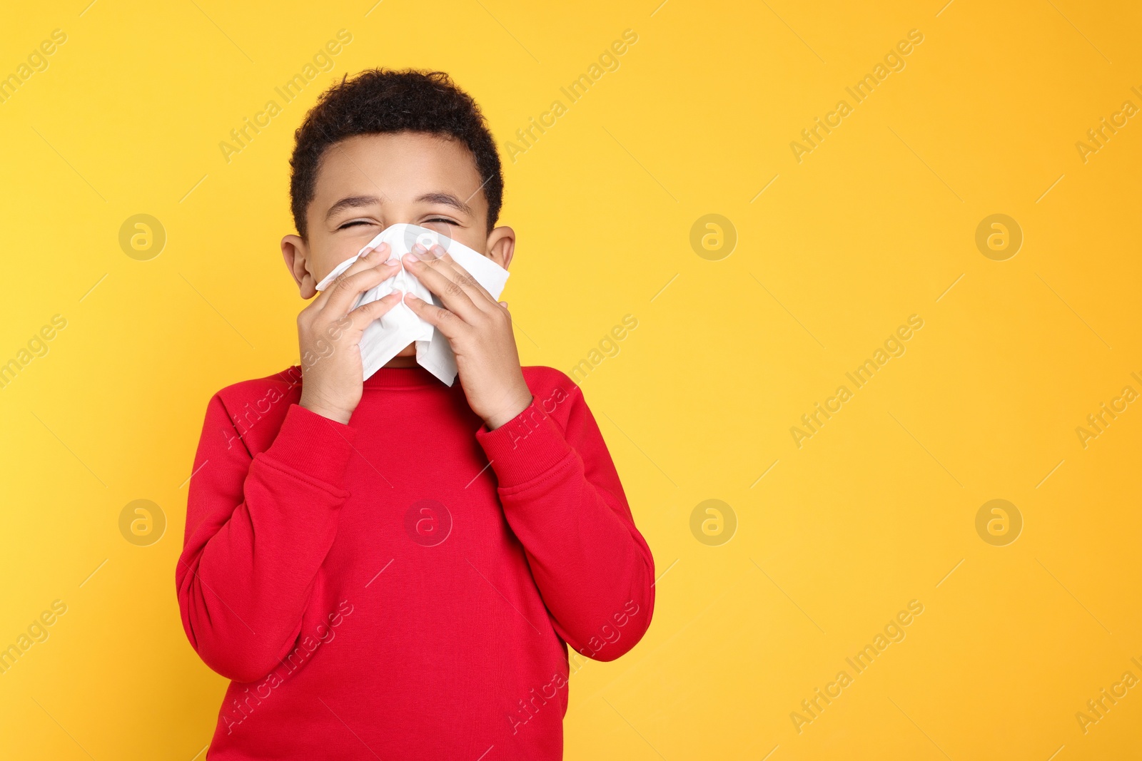 Photo of African-American boy blowing nose in tissue on yellow background, space for text. Cold symptoms