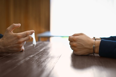 Photo of Police officer interrogating drug dealer in handcuffs at desk indoors. Criminal law