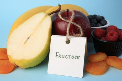 Photo of Card with word Fructose, delicious ripe fruits, berries and dried apricots on light blue background, closeup