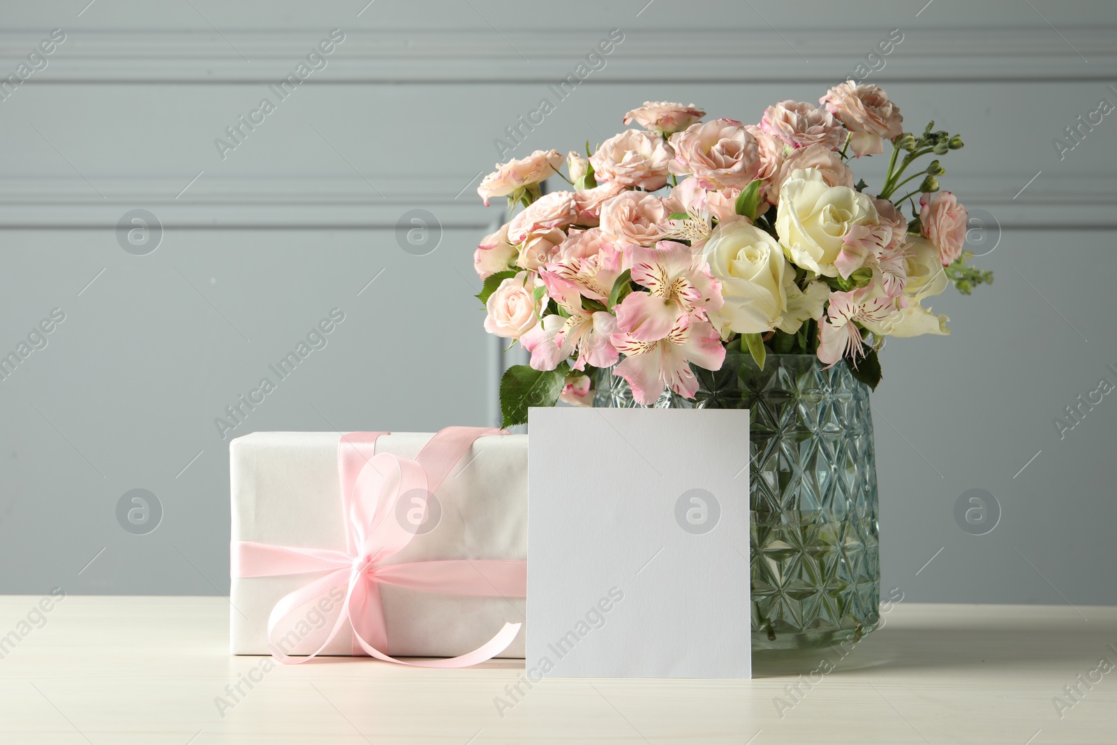 Photo of Happy Mother's Day. Gift box, blank card and bouquet of beautiful flowers in vase on white table near grey wall