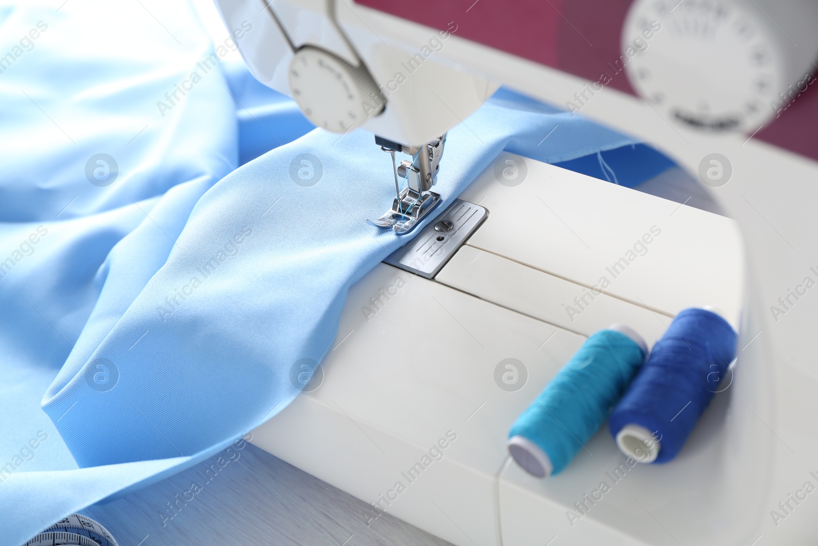 Photo of White sewing machine with light blue fabric and spools of threads on table, closeup