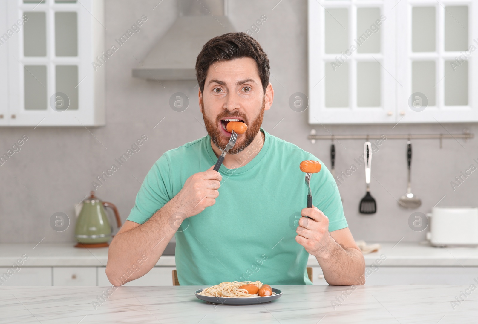 Photo of Man eating sausage and pasta at table in kitchen