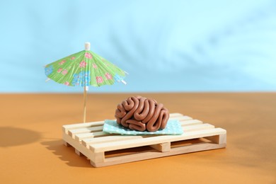 Brain made of plasticine on mini wooden sunbed under umbrella against color background