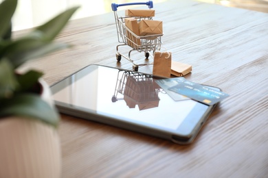 Photo of Internet shopping. Small cart with boxes, bags and credit cards near modern tablet on wooden table