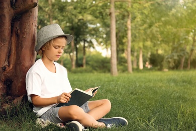 Photo of Cute little boy reading book on green grass near tree in park
