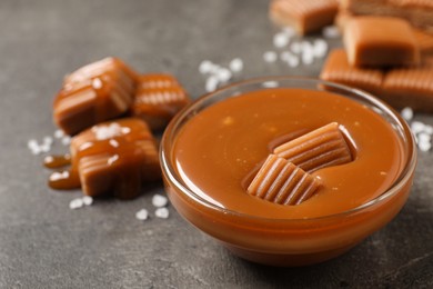 Photo of Tasty salted caramel with candies in glass bowl on grey table, closeup