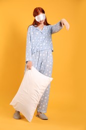 Young woman wearing pajamas and slippers with pillow in sleepwalking state on yellow background