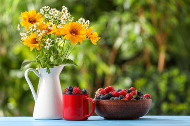 Photo of Different fresh ripe berries and beautiful flowers on light blue table outdoors