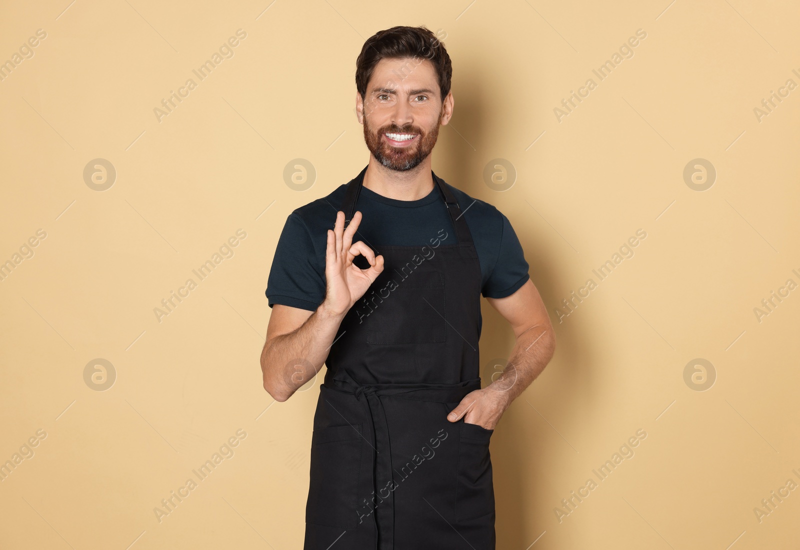 Photo of Smiling hairdresser wearing apron showing ok gesture on light brown background