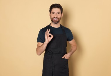 Photo of Smiling hairdresser wearing apron showing ok gesture on light brown background
