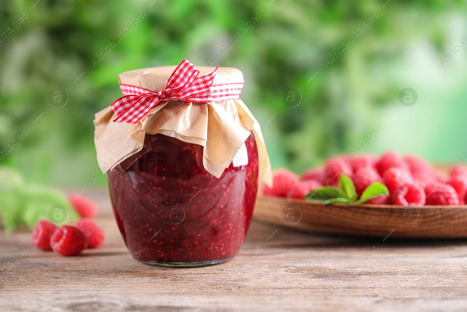 Photo of Glass jar of sweet jam with ripe raspberries on wooden table against blurred background. Space for text