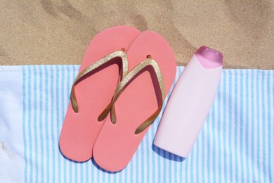Striped towel with bottle of sunblock and flip flops on sandy beach, flat lay