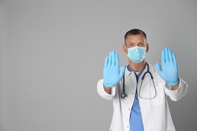 Photo of Doctor in protective mask showing stop gesture on grey background, space for text. Prevent spreading of coronavirus